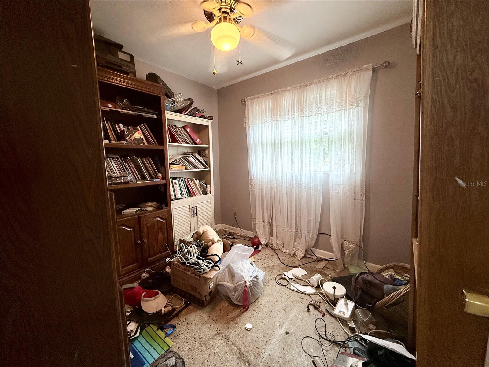 Cluttered room with book-filled shelves, lace curtains, and belongings scattered across the floor.