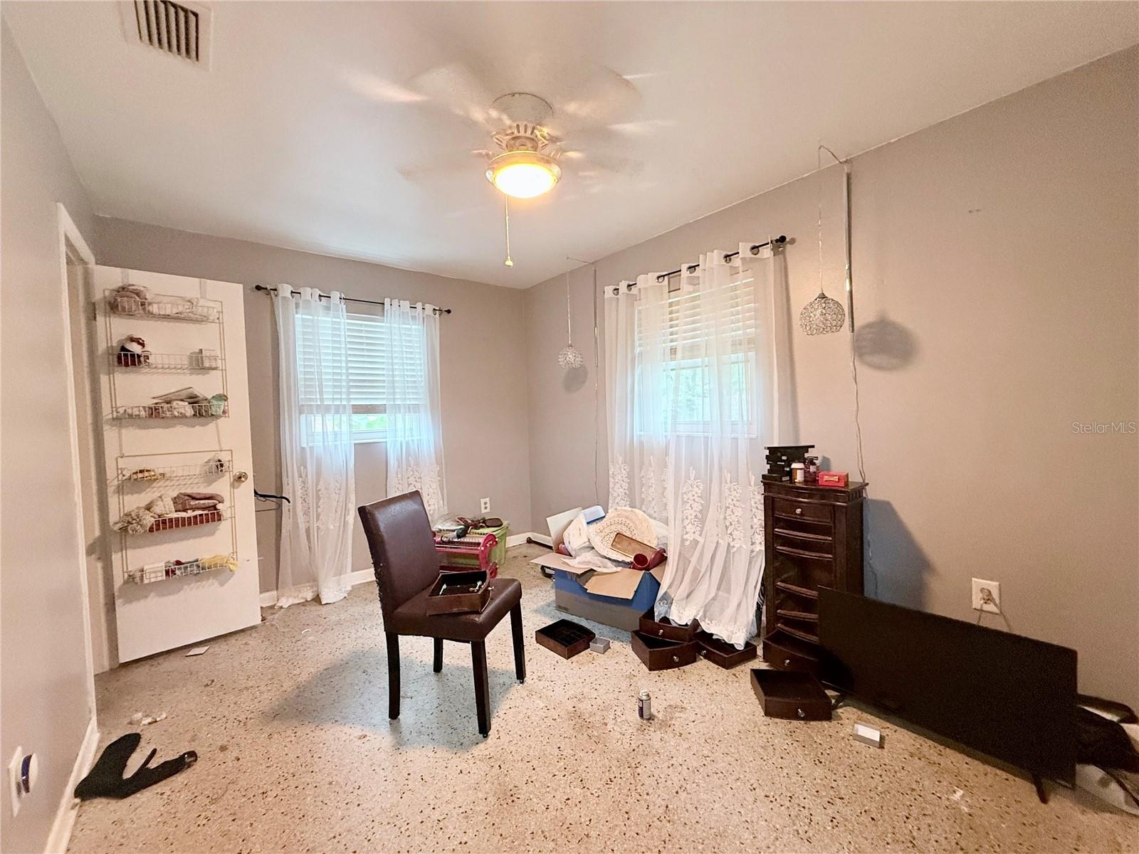 Interior room with a ceiling fan, two curtained windows, terrazzo flooring, and belongings scattered around.