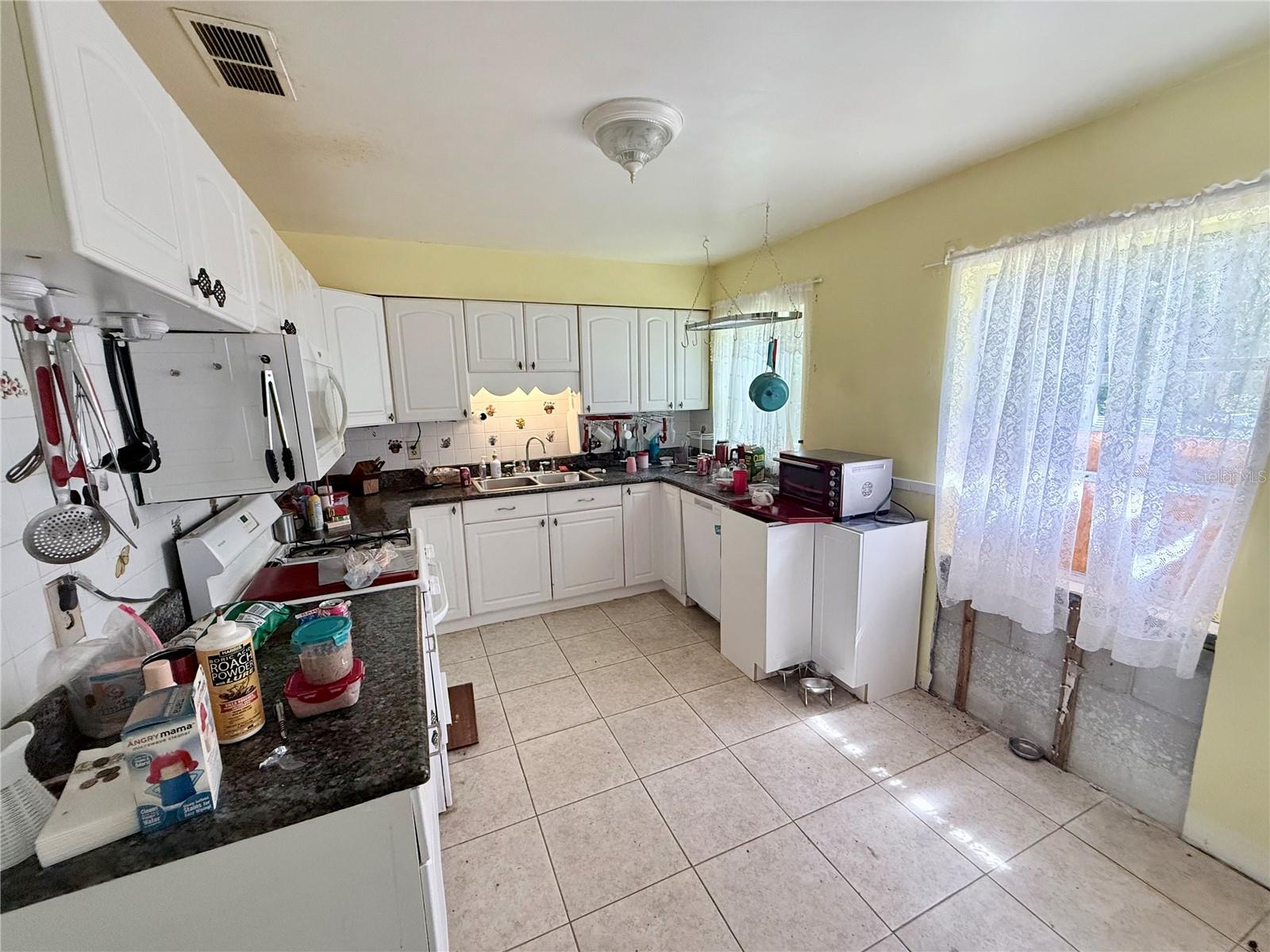 Kitchen with white cabinets, dark counters, tiled floors, and lace curtains over the window and sliding door.