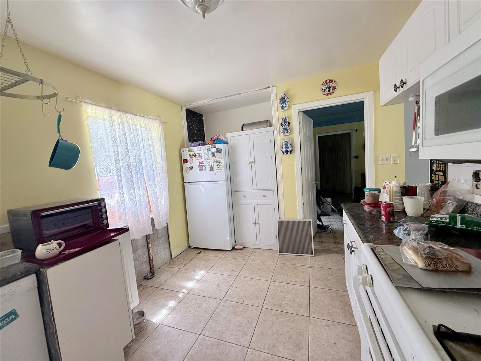 Kitchen with yellow walls, lace-curtained window, magnet-covered refrigerator, and crowded countertop.
