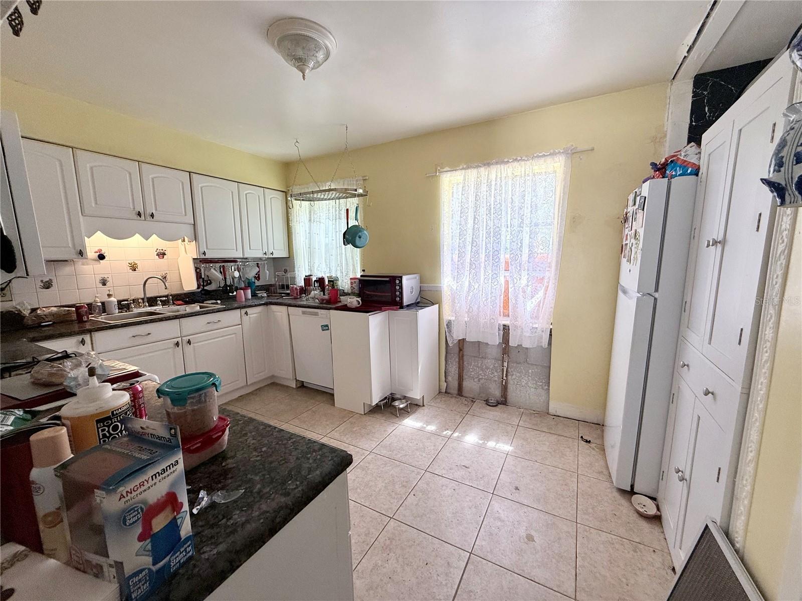 Cluttered kitchen with white cabinets, dark countertops, and lace curtains over a glass door.