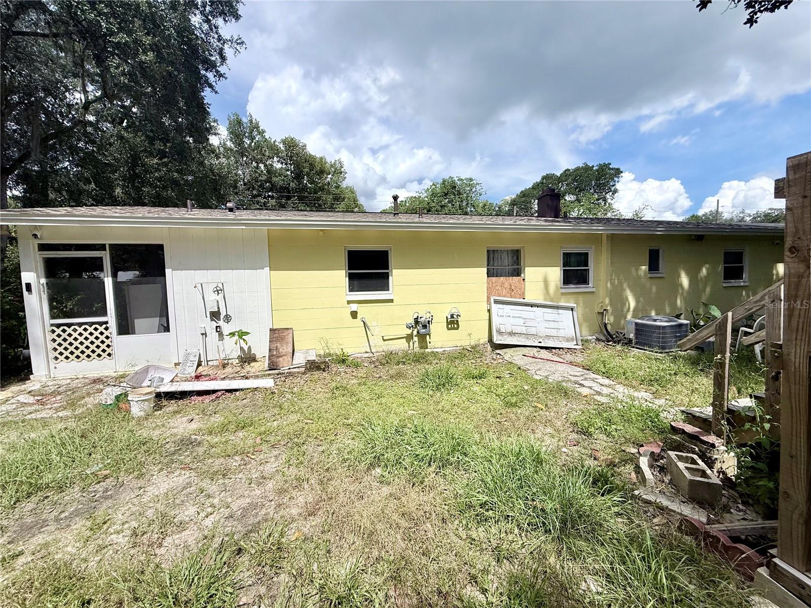 Rear of a yellow single-story house with an overgrown yard, scattered debris, and a boarded window.