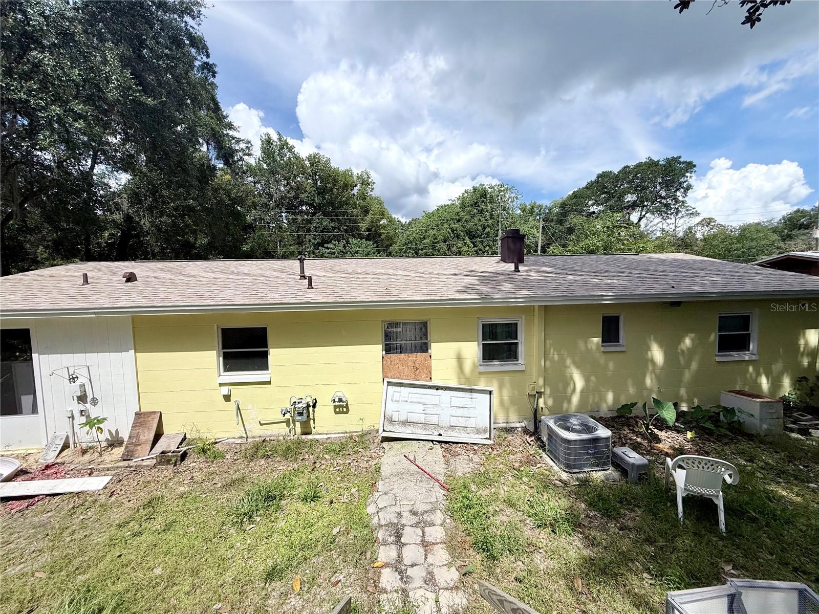 Rear exterior of a yellow single-story house with a boarded window and a stone path through the yard.