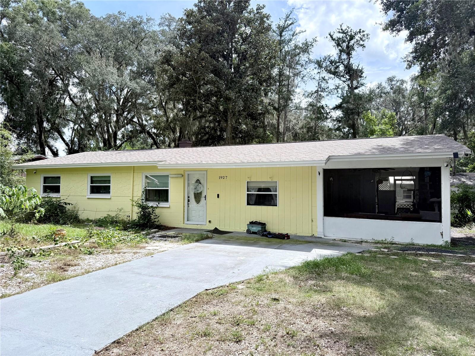 Pale yellow single-story home with a wide driveway and screened porch beneath mature trees.
