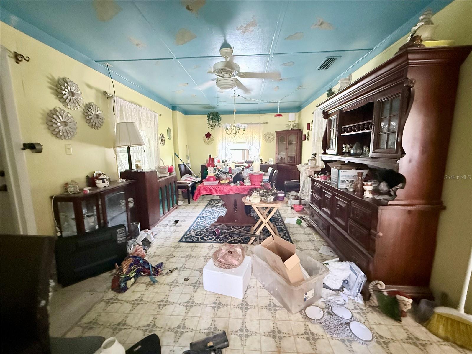 Cluttered dining room with dark wood cabinets, patterned tile floors, and peeling paint on the blue ceiling.