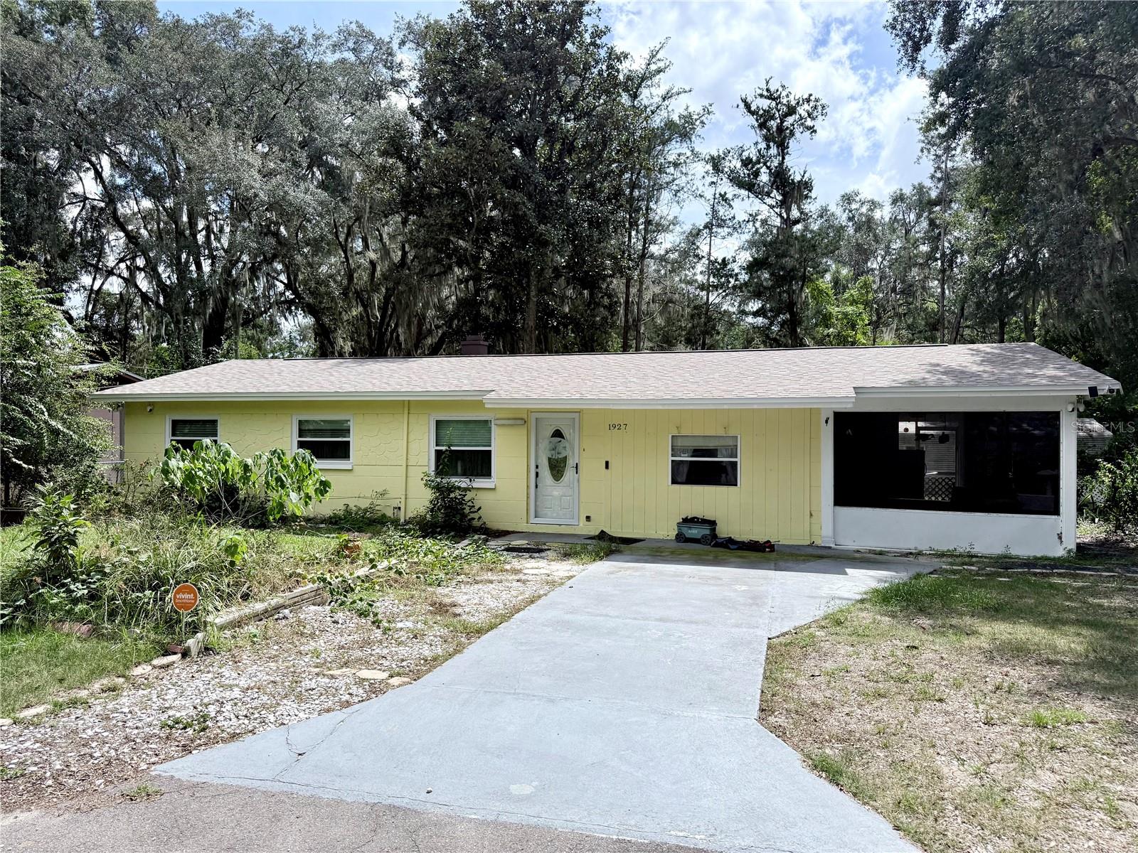 Single-story yellow house with a screened porch and concrete driveway beneath mature trees.