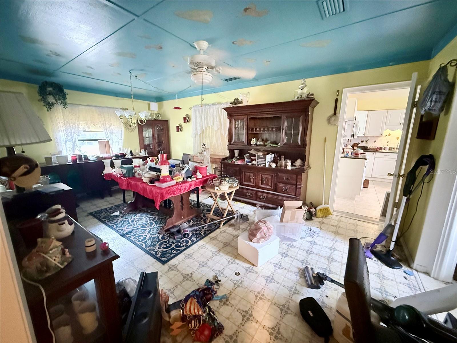 Cluttered living and dining room with blue ceiling, dark wood china cabinet, and doorway to a white kitchen.