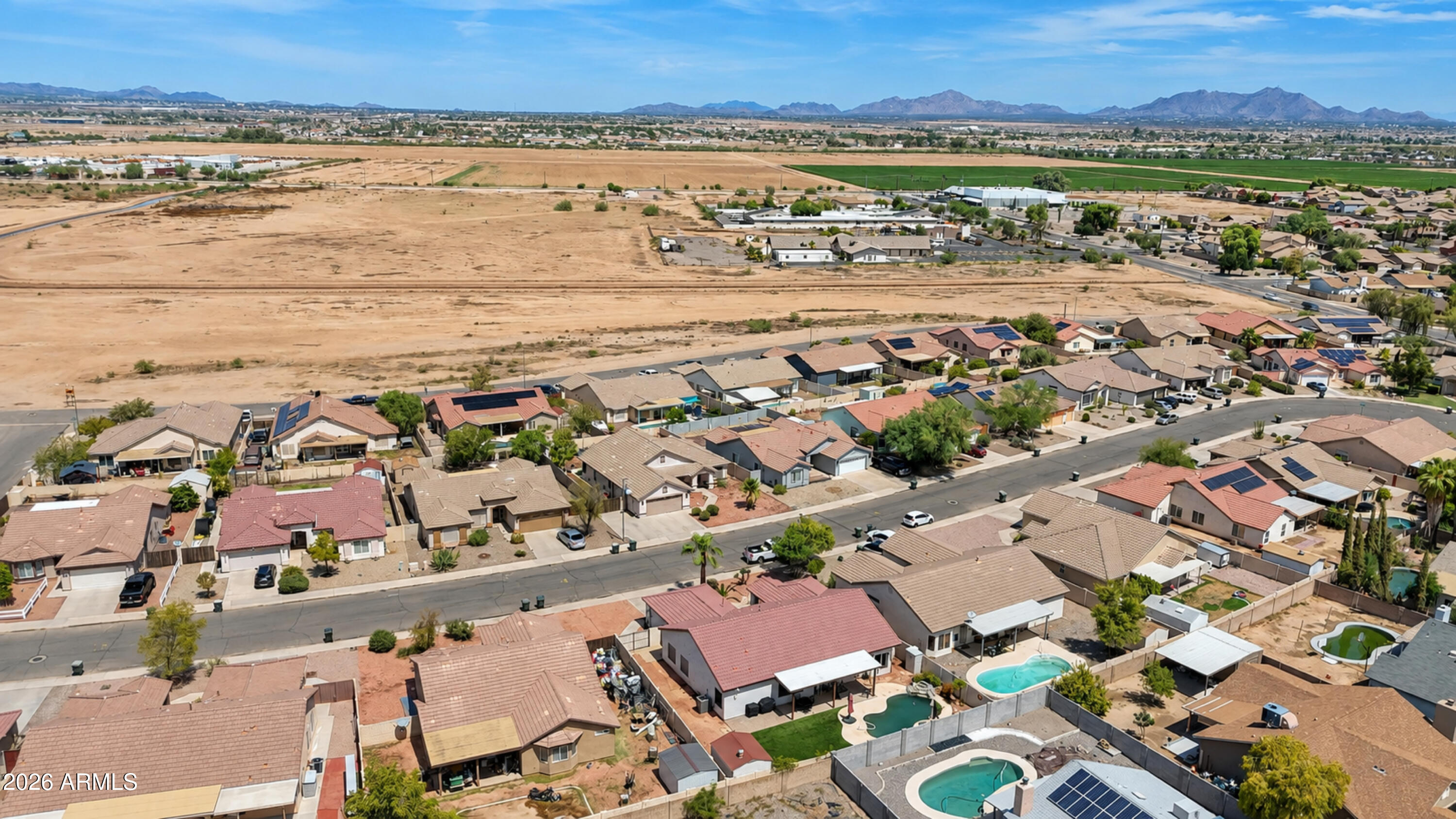 Aerial view of homes and backyard pools beside a large undeveloped desert tract, with mountains in the distance.