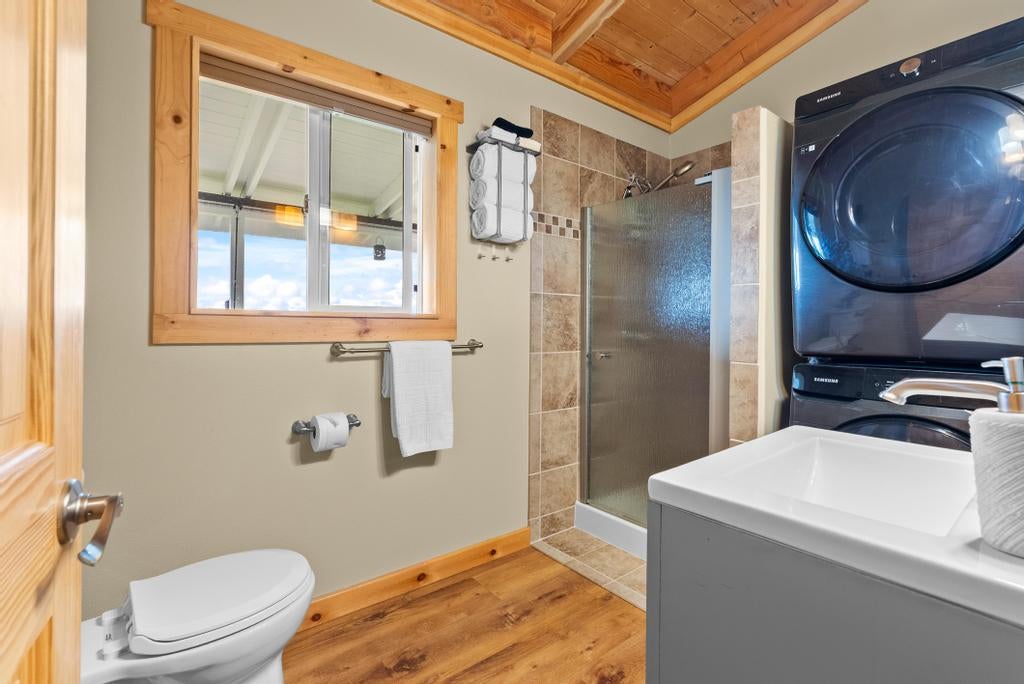 Bathroom with a tiled glass shower, stacked washer and dryer, and warm wood flooring and ceiling.