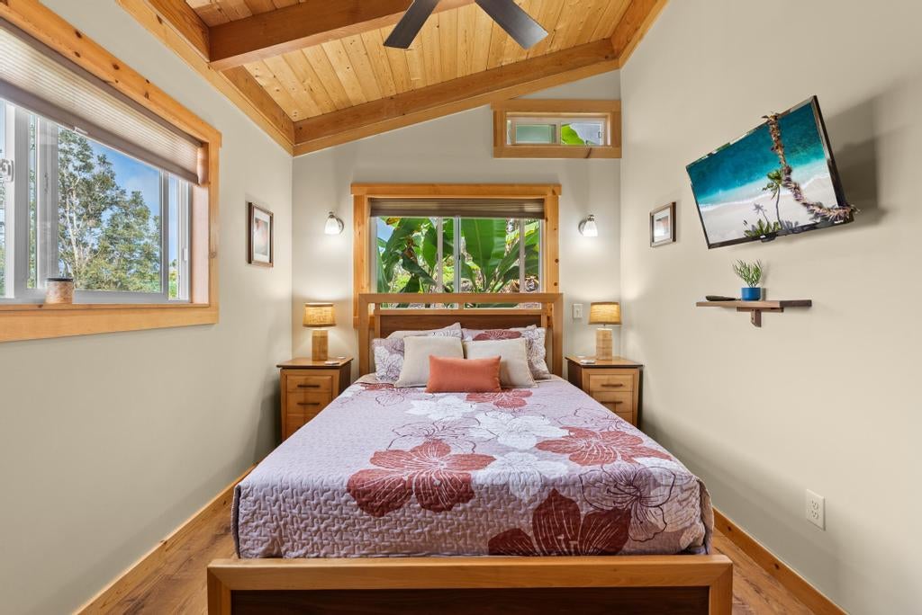 Bedroom with a floral quilt, vaulted wood ceiling, and windows overlooking tropical foliage.