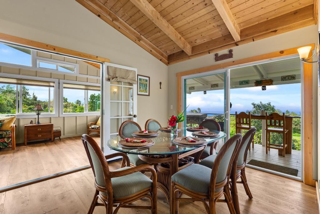Dining room with a glass-top table, vaulted wood ceiling, and ocean view through open sliding doors.