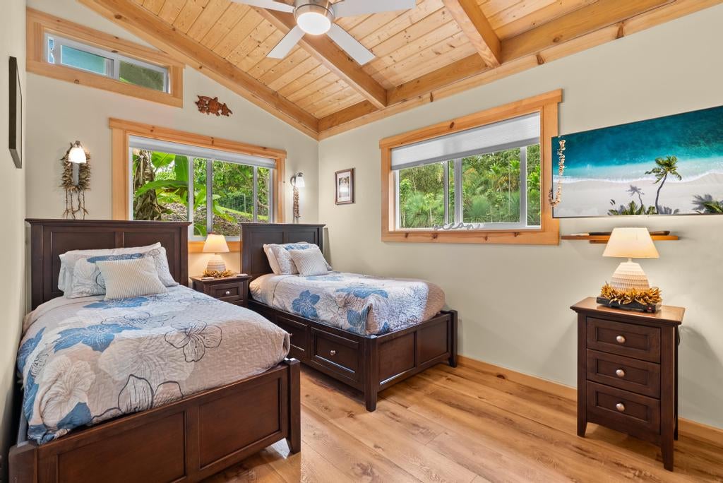 Bedroom with two dark wood beds beneath a vaulted wood ceiling and windows overlooking tropical greenery.