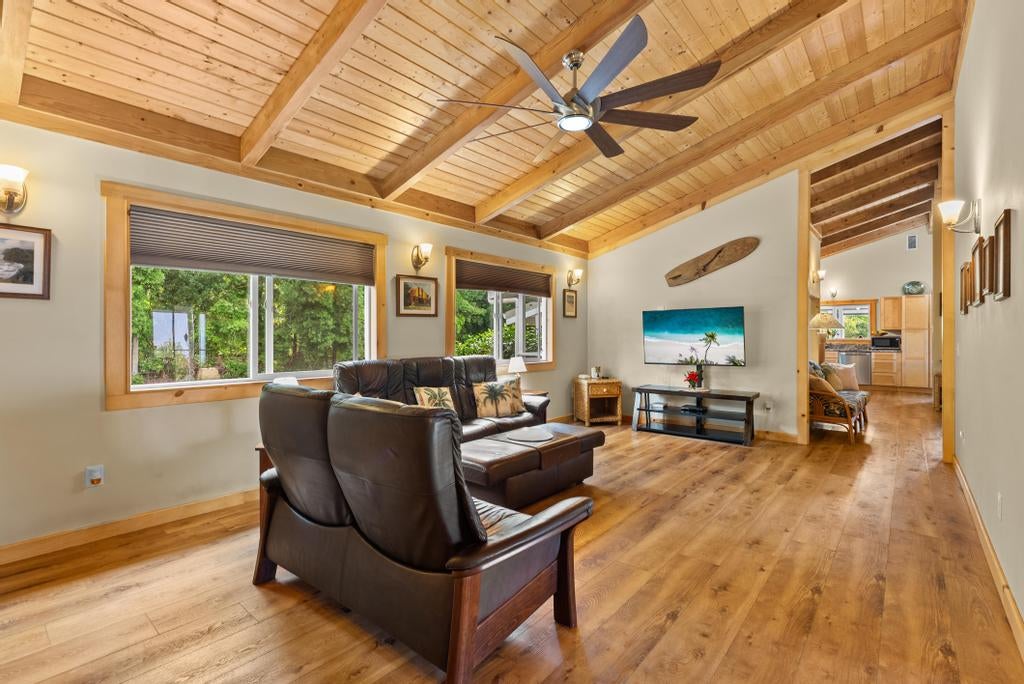 Living room with vaulted wood ceiling, exposed beams, leather seating, and windows overlooking greenery.