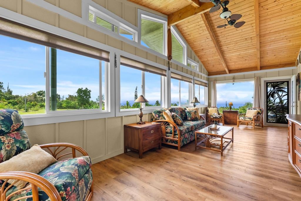 Living room with a vaulted wood ceiling, floral seating, and wide windows overlooking greenery and ocean.