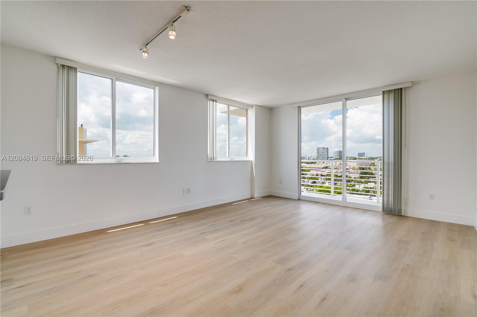 Vacant condo living room with light wood floors, large windows, and sliding doors to a balcony.