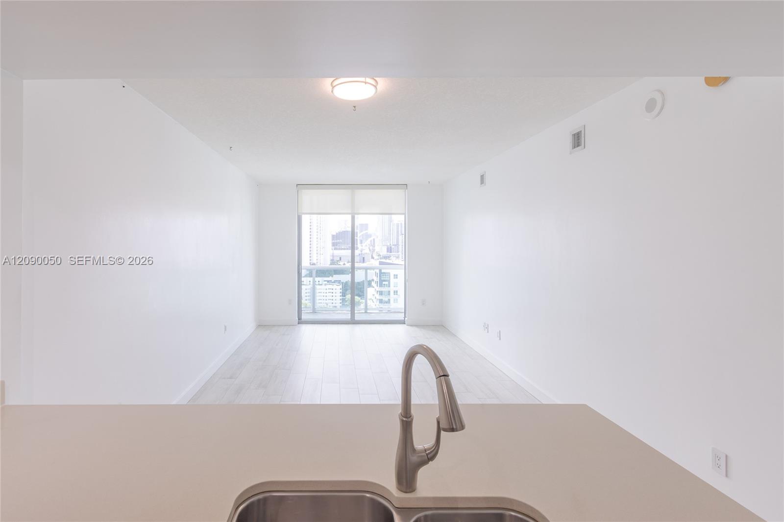Vacant condo living area with pale floors, a balcony, and Miami buildings visible through sliding glass doors.