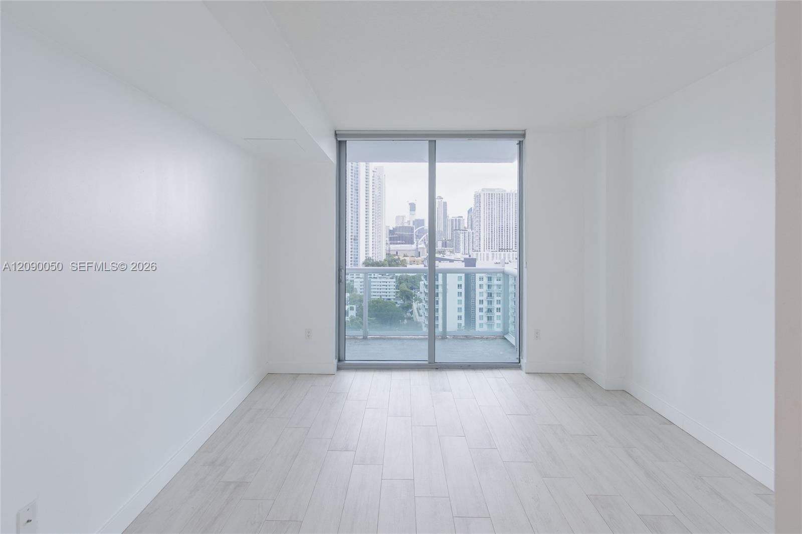 Empty bright room with pale plank flooring and sliding doors opening to a balcony with city views.