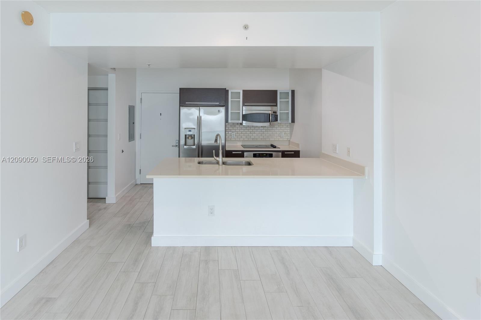 Vacant condo kitchen with a white peninsula, stainless steel appliances, dark cabinets, and pale plank flooring.