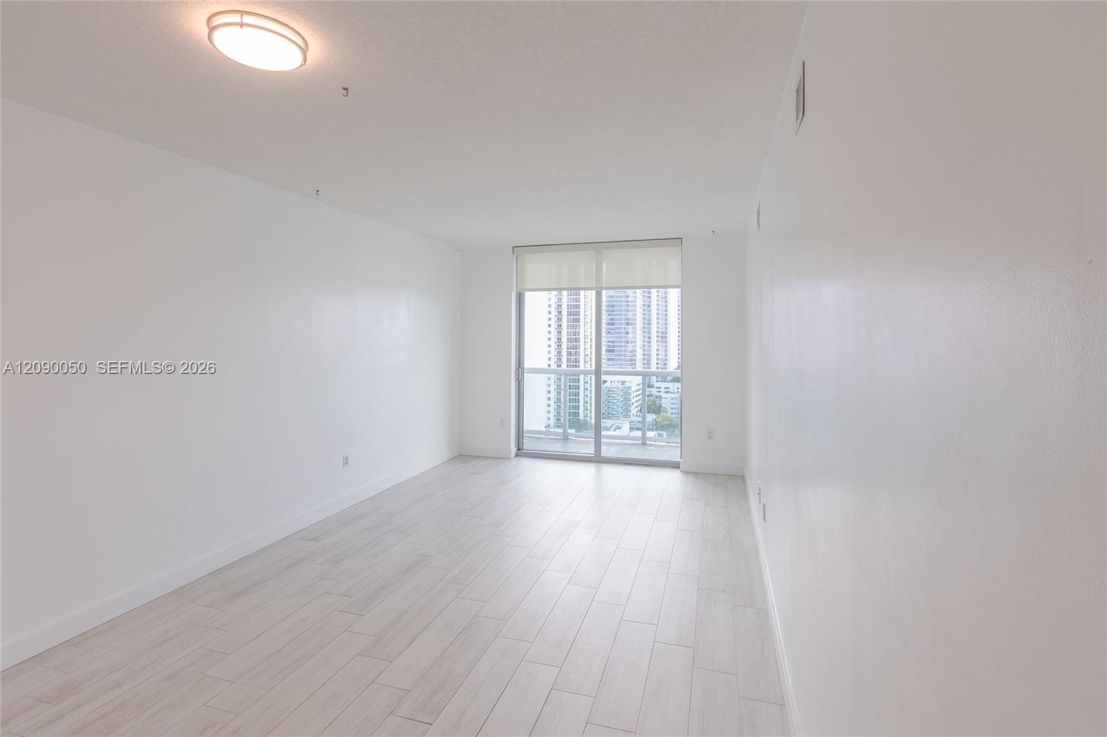 Empty condo room with pale tile floors, sliding balcony doors, and a view of nearby high-rises.