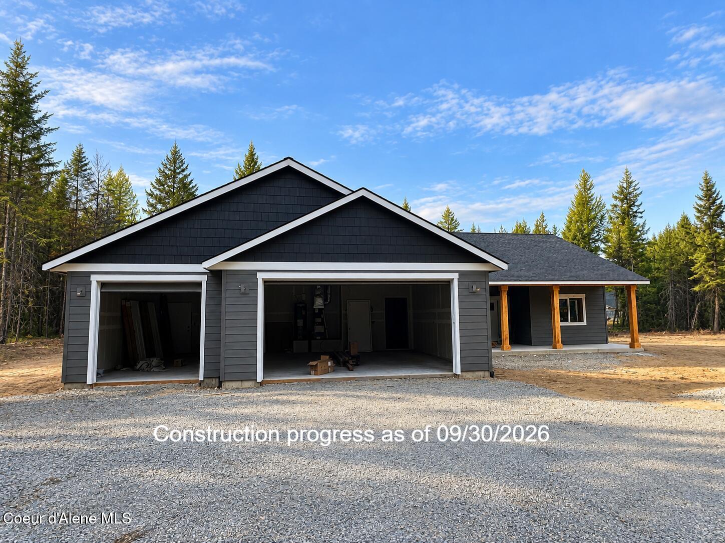 Dark gray home under construction with two open garage bays and a covered porch among evergreen trees.