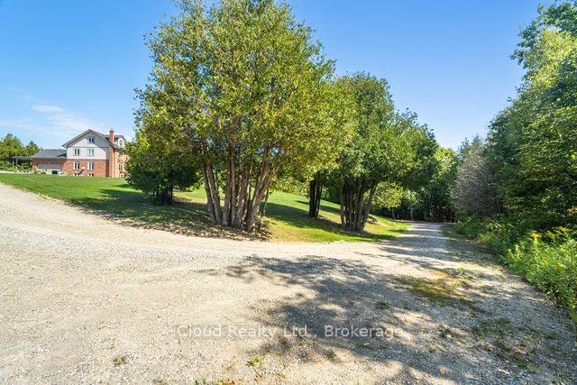Rural house beyond a gravel driveway, framed by mature leafy trees.
