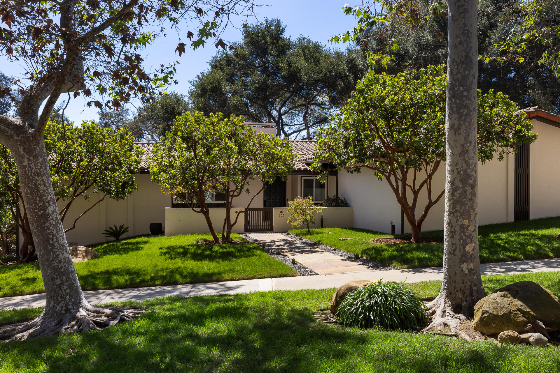 Single-story stucco home with a tiled roof, shaded entry path, mature trees, and landscaped lawn.