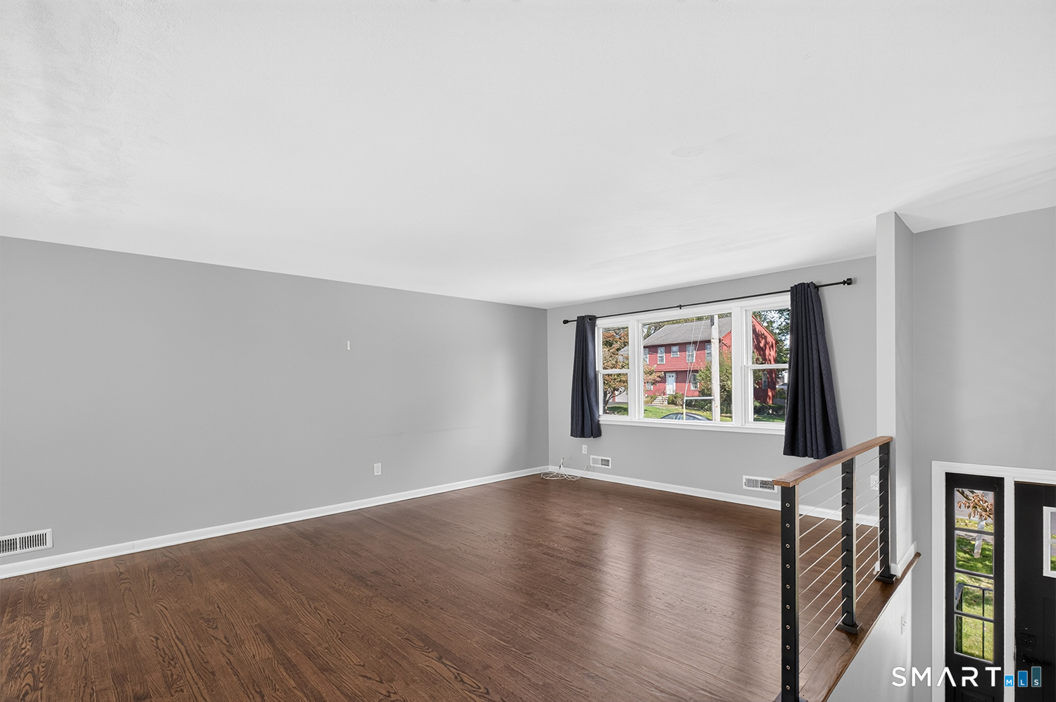 Vacant living room with dark hardwood floors, a large curtained window, and a metal stair railing.