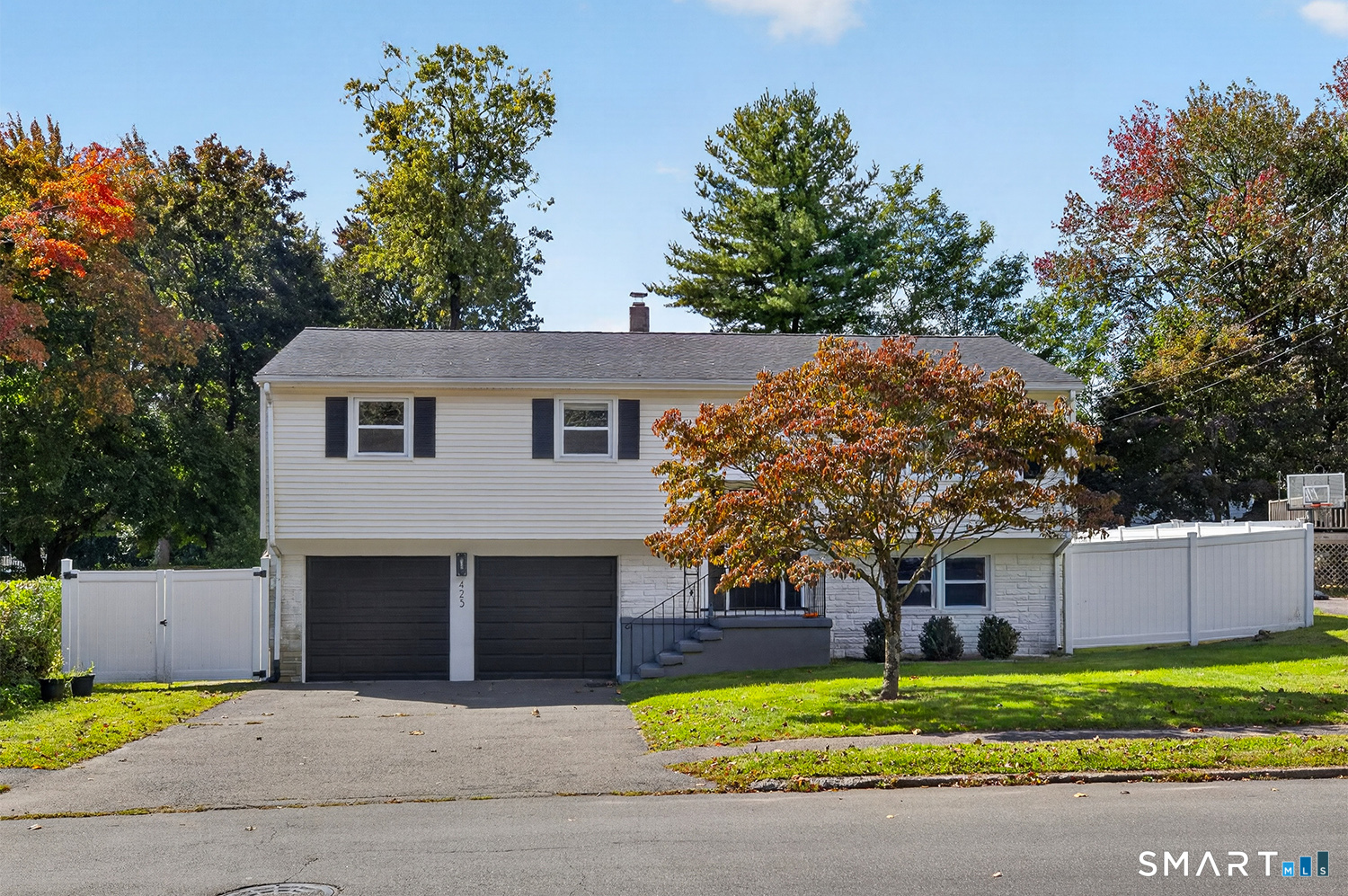 White split-level home with two garage doors, white fencing, and an autumn tree in front.
