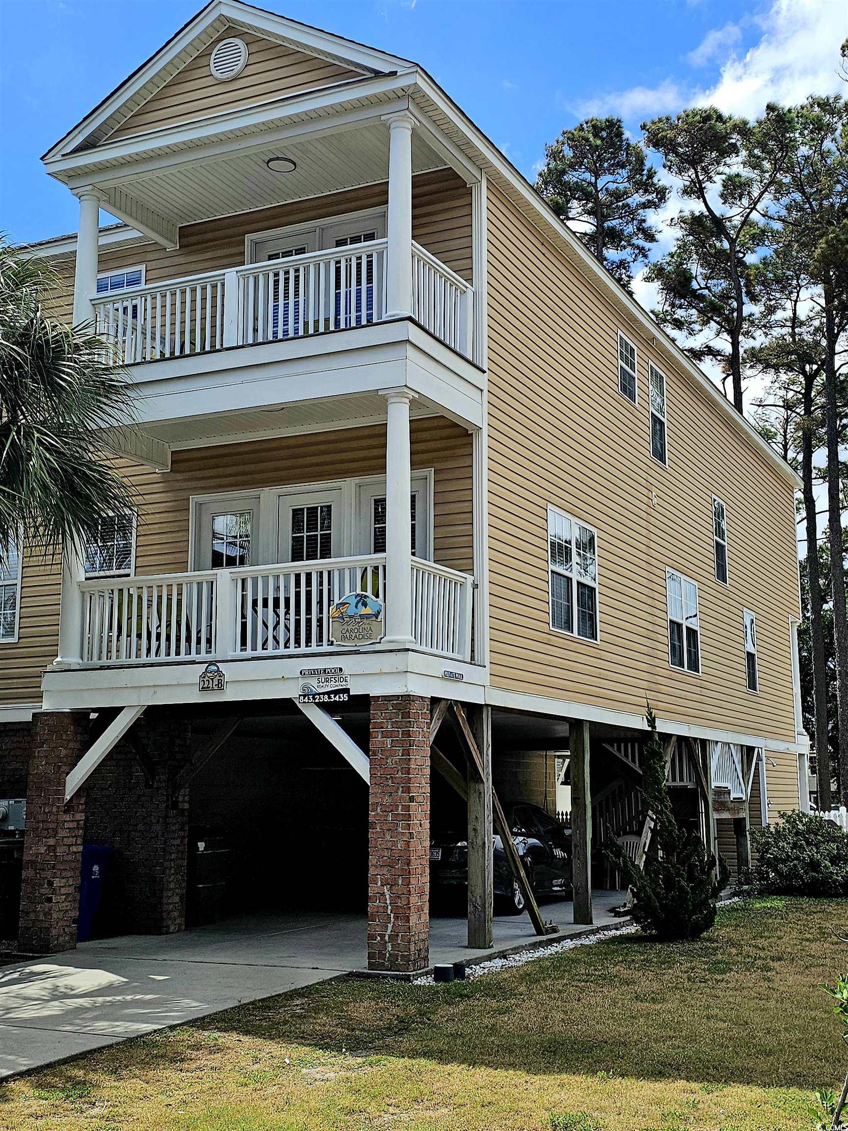Raised tan home with two white-railed balconies and brick supports beneath the parking level.