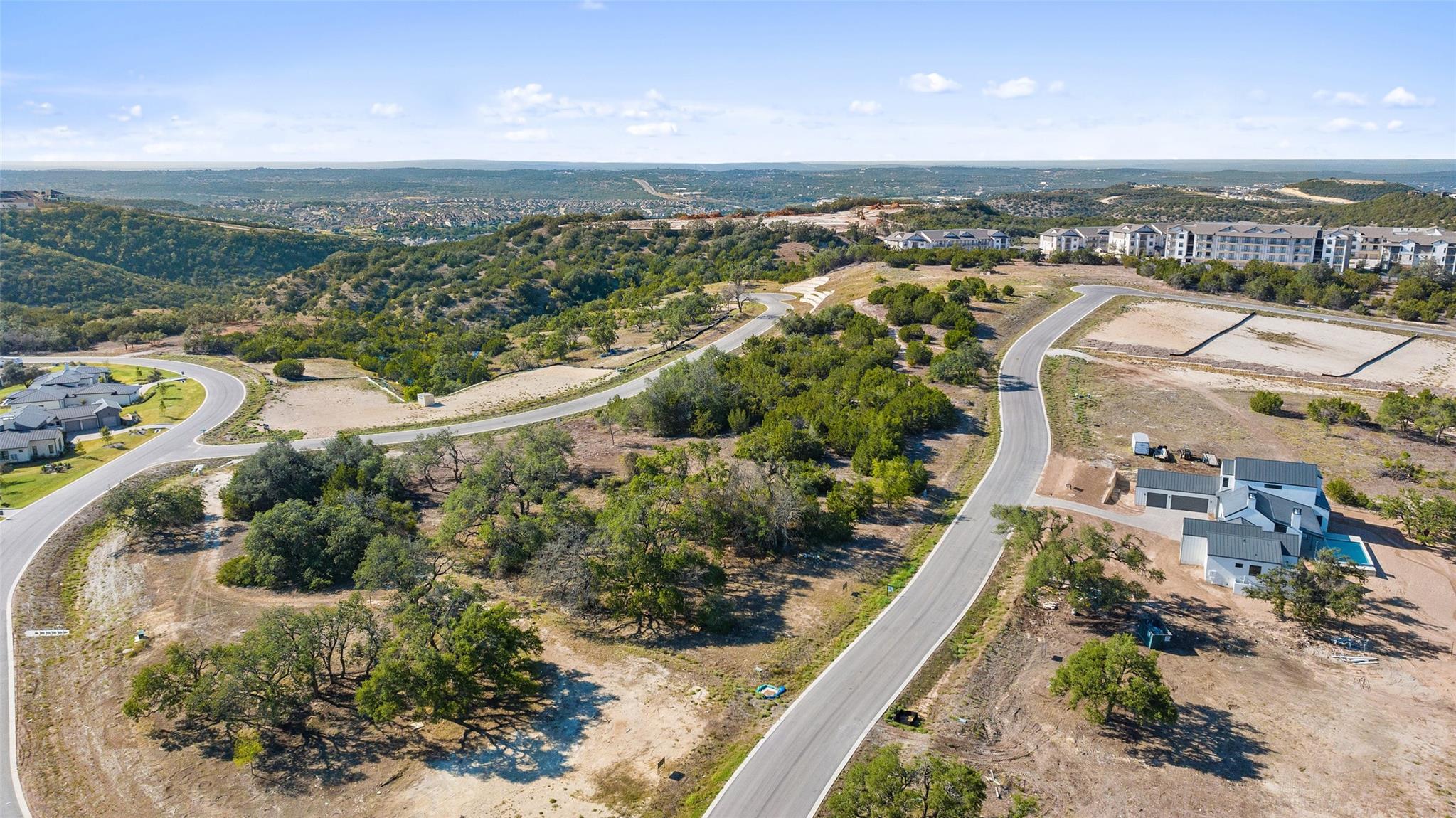 Aerial view of undeveloped lots and curving roads, with homes, apartments, and rolling hills in the distance.