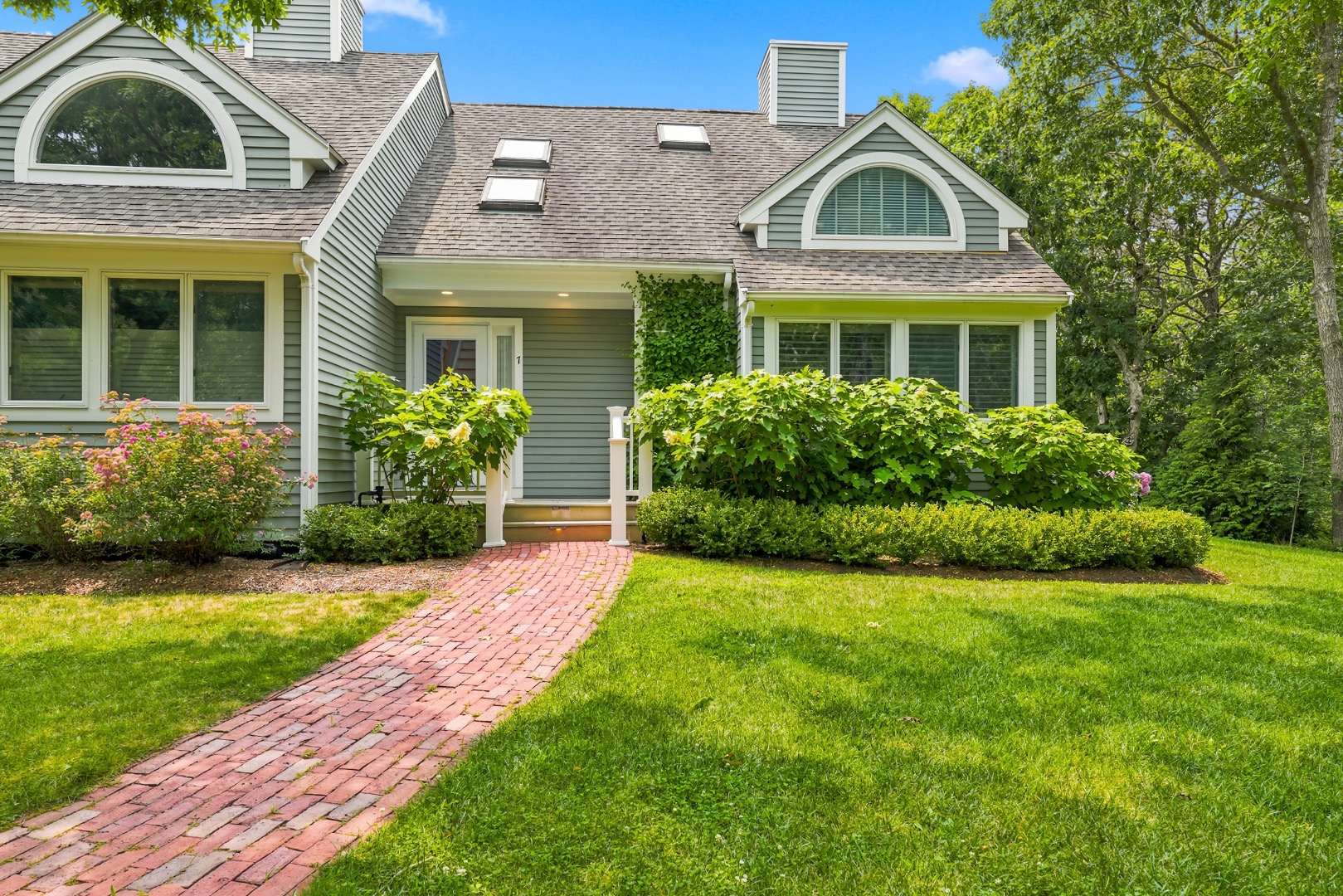 Gray-green house with arched dormers, a brick walkway, and lush garden landscaping.