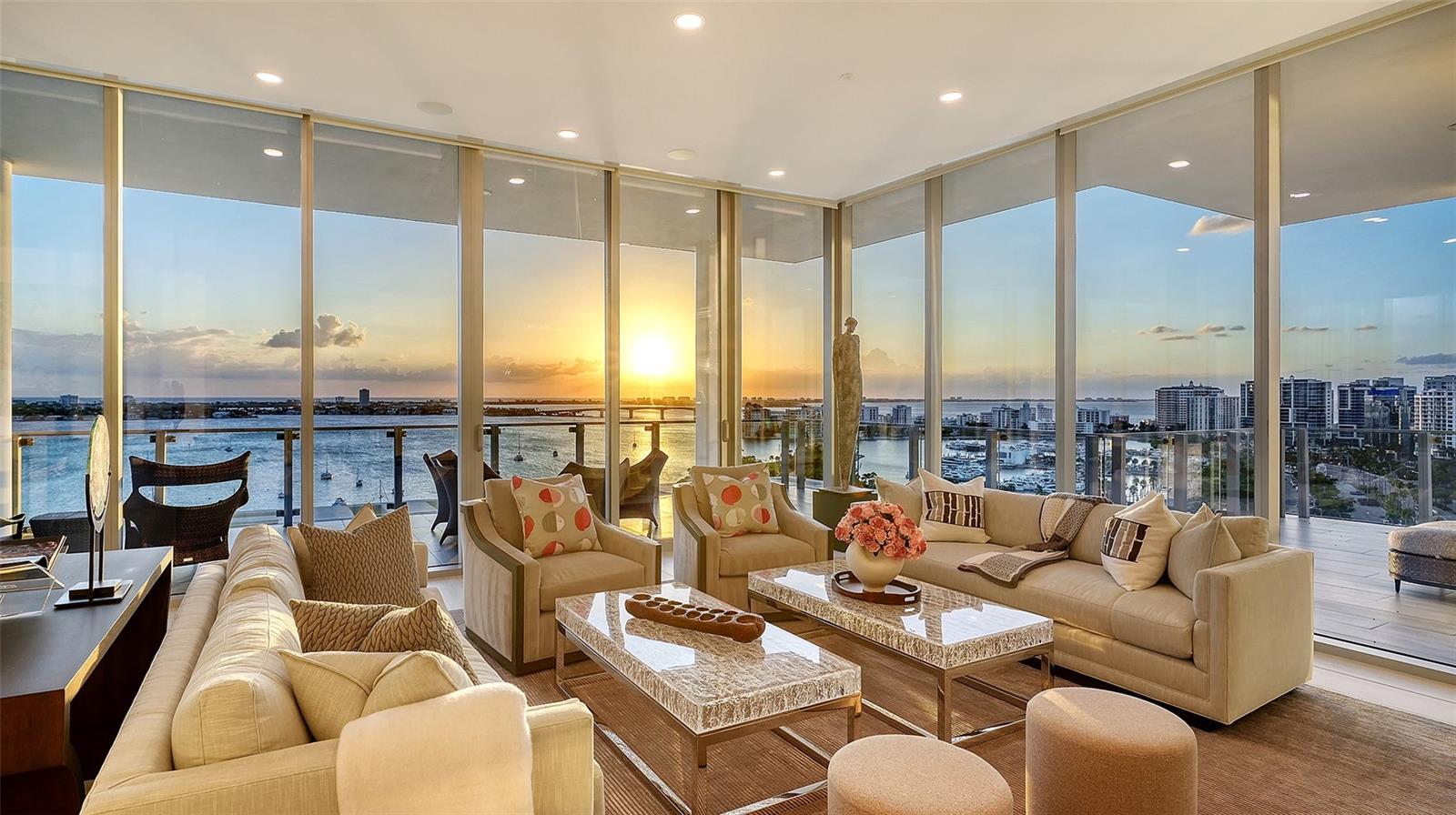 Furnished living room with floor-to-ceiling windows overlooking Sarasota Bay and downtown at sunset.