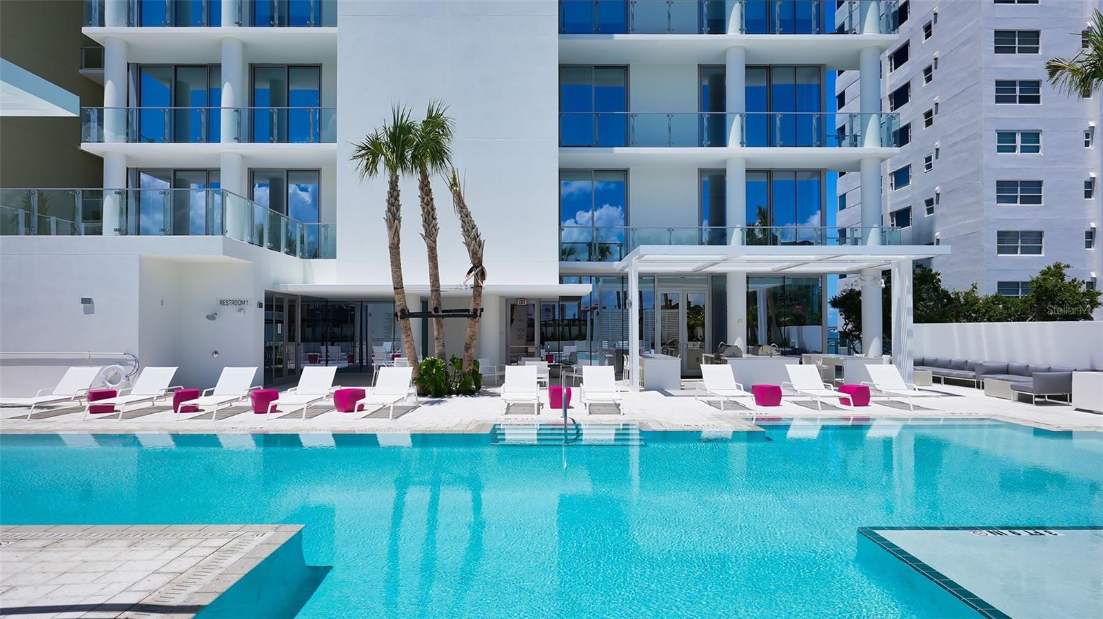 Condominium pool with white loungers, pink side tables, and glass balconies.