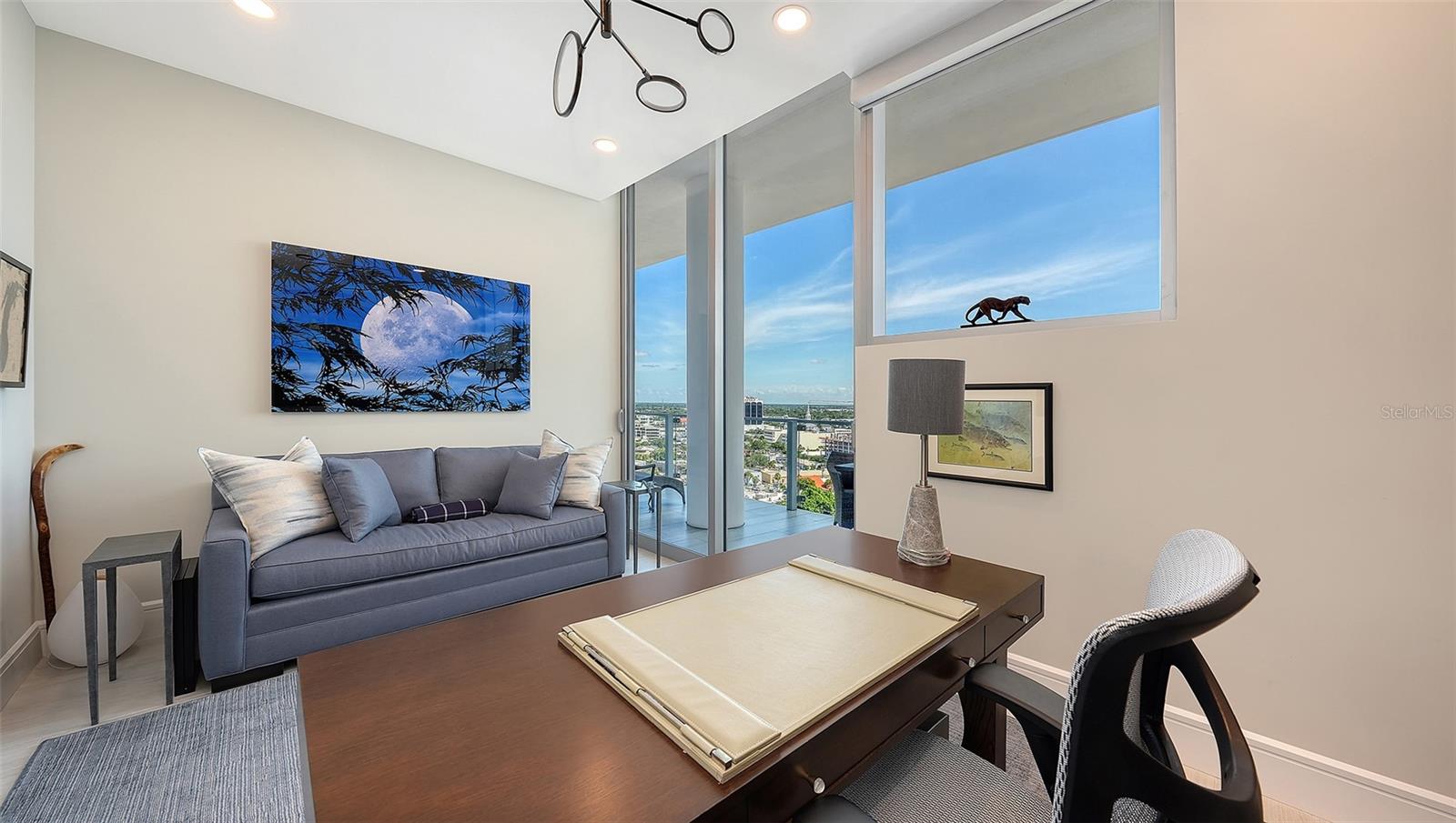 Furnished condo office with a gray sofa, dark wood desk, and expansive windows overlooking the city.