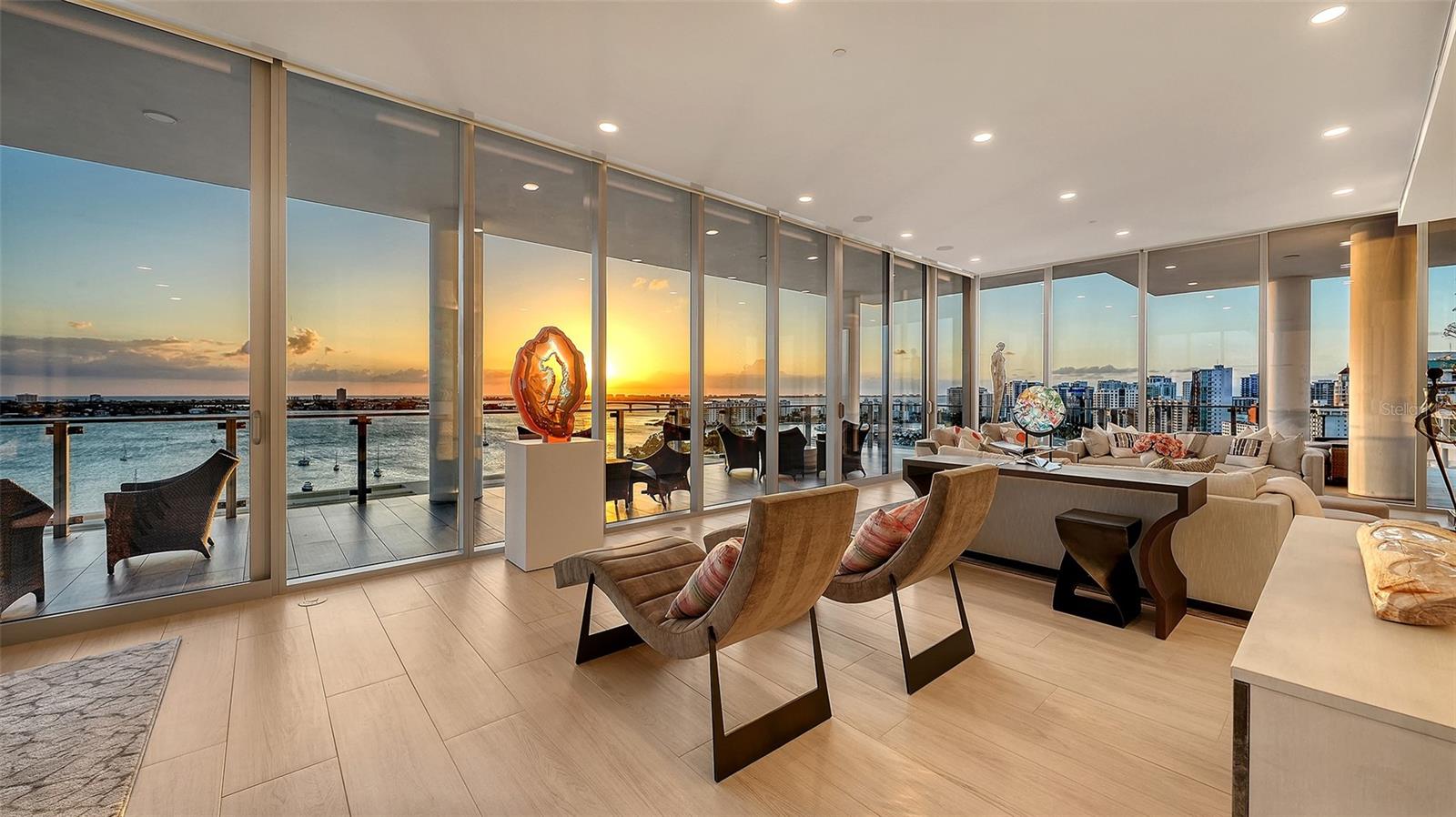 Furnished condo living room with floor-to-ceiling windows overlooking Sarasota Bay at sunset.