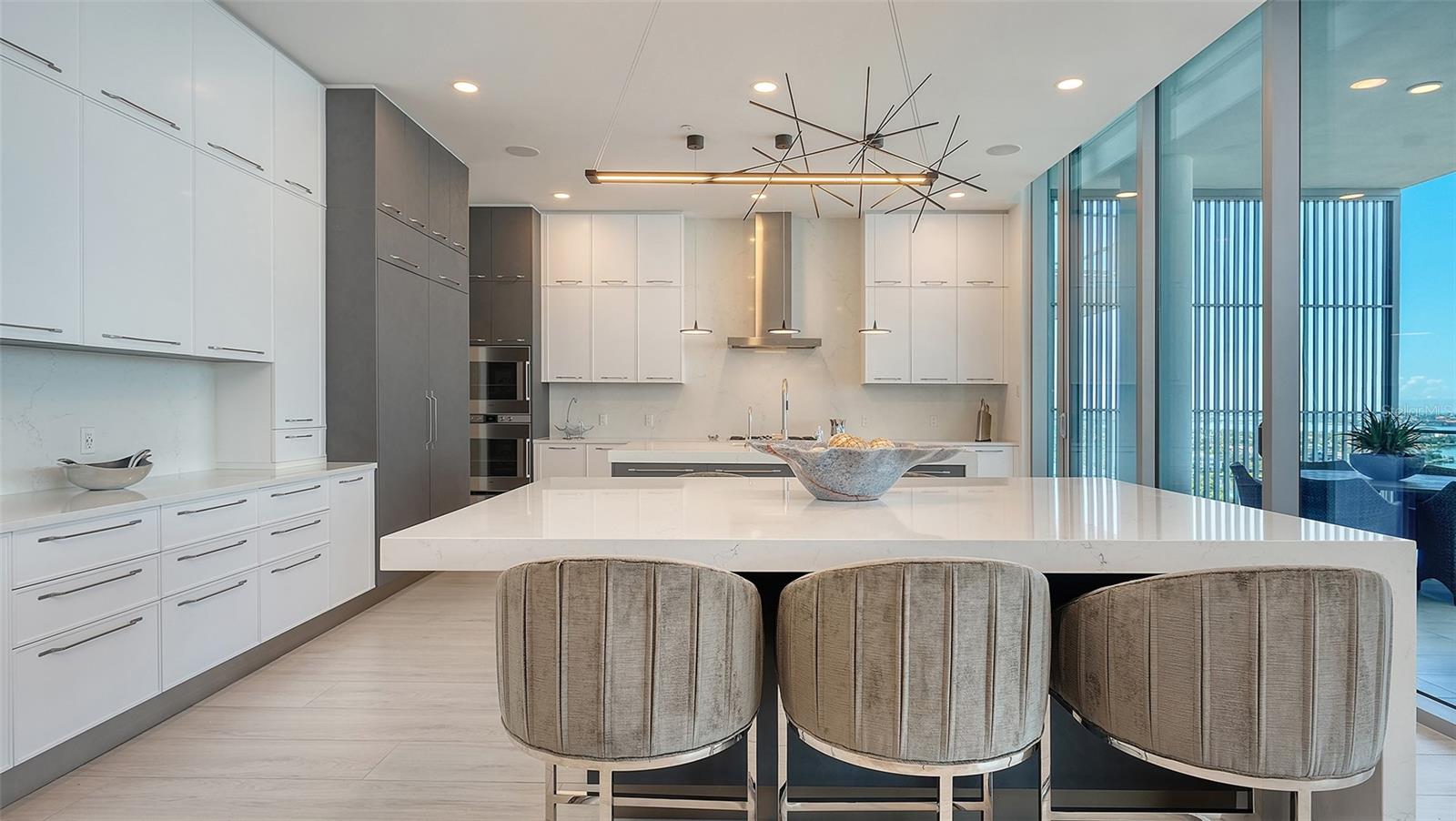 Modern kitchen with a large island, three stools, and floor-to-ceiling windows overlooking water.