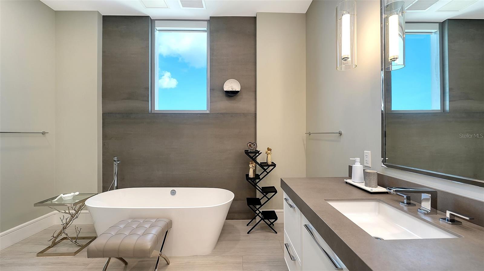 Modern bathroom with a freestanding soaking tub, gray tile, and high windows showing blue sky.