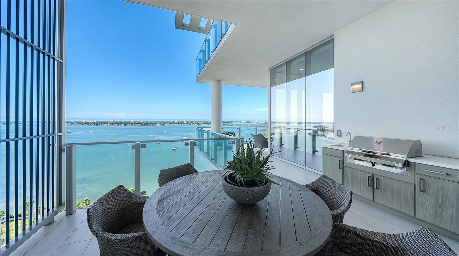 Condo balcony with an outdoor dining set and grill overlooking a sailboat-filled bay.