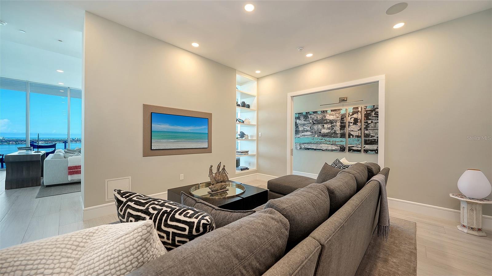 Condo living room with a gray sectional and built-in shelves, opening to water views through tall windows.