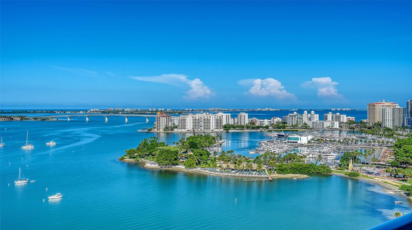 Aerial view of Sarasota Bay with boats, a marina, a bridge, and waterfront buildings.