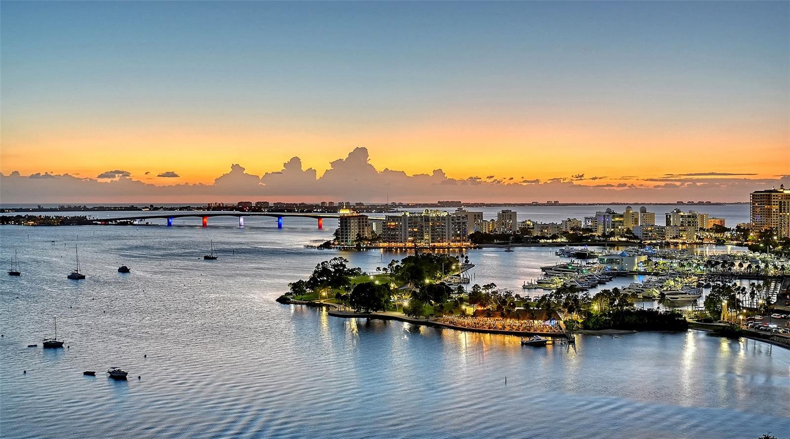 Sarasota Bay and marina at sunset, with sailboats, waterfront buildings, and an illuminated bridge.