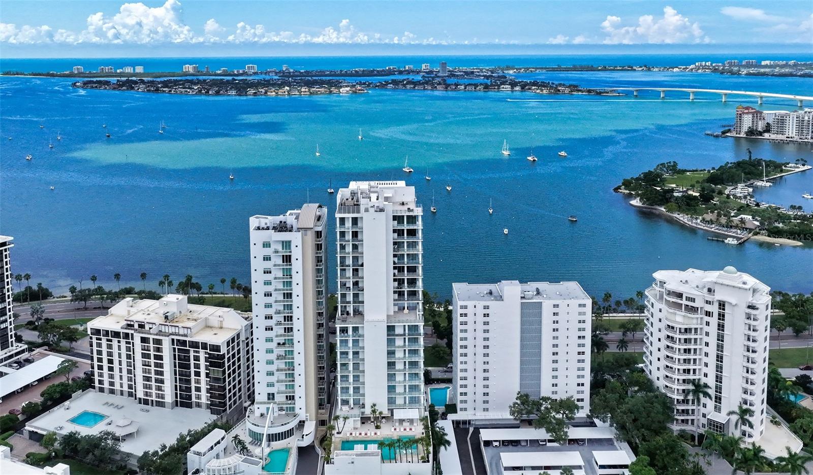 Waterfront condominium towers overlooking a blue bay, with swimming pools, sailboats, and a distant bridge.