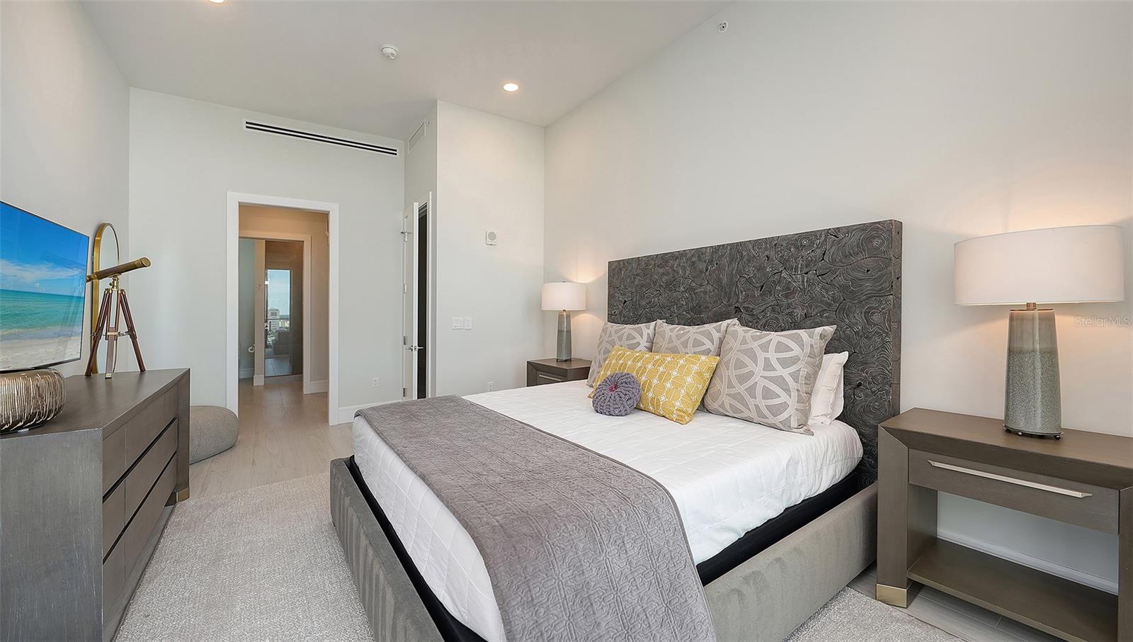 Staged bedroom with a large bed, dark textured headboard, and matching gray bedside tables.