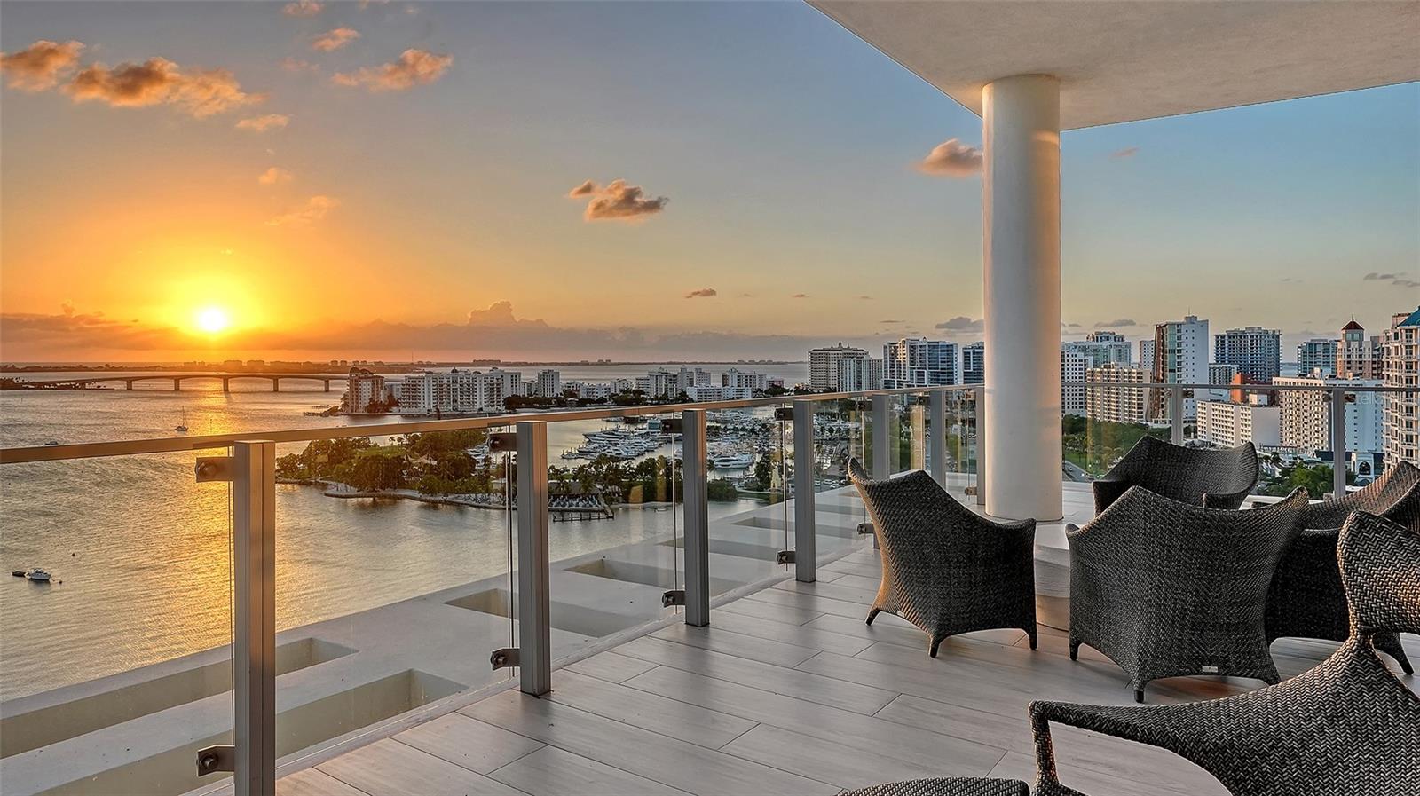 High-rise balcony with woven chairs overlooking a marina and waterfront skyline at sunset.