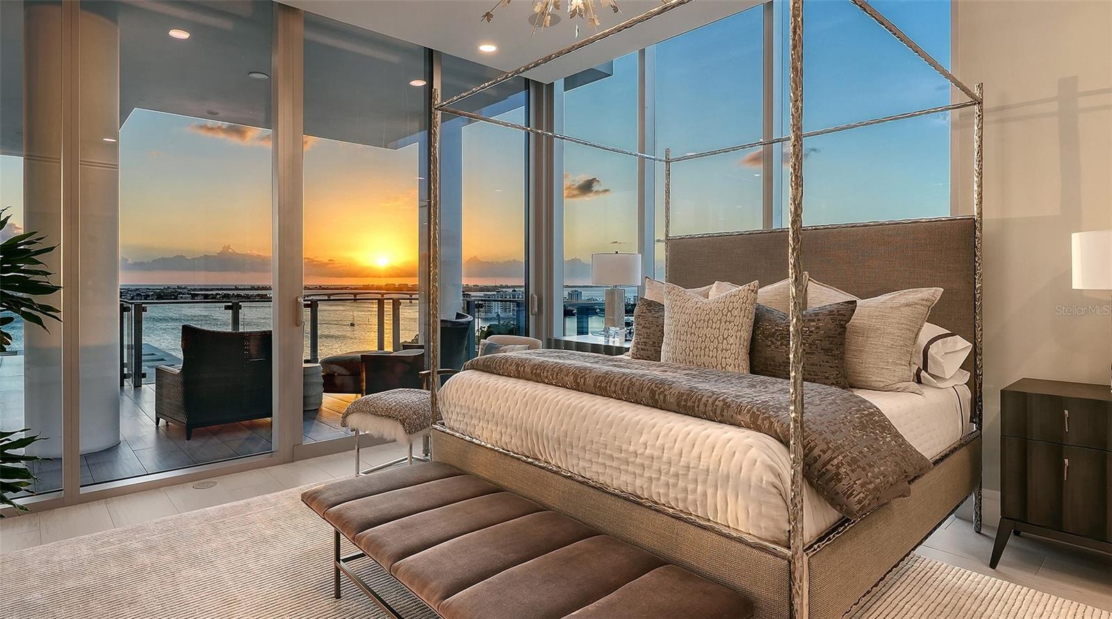 Bedroom with a canopy bed and floor-to-ceiling windows overlooking the water at sunset.