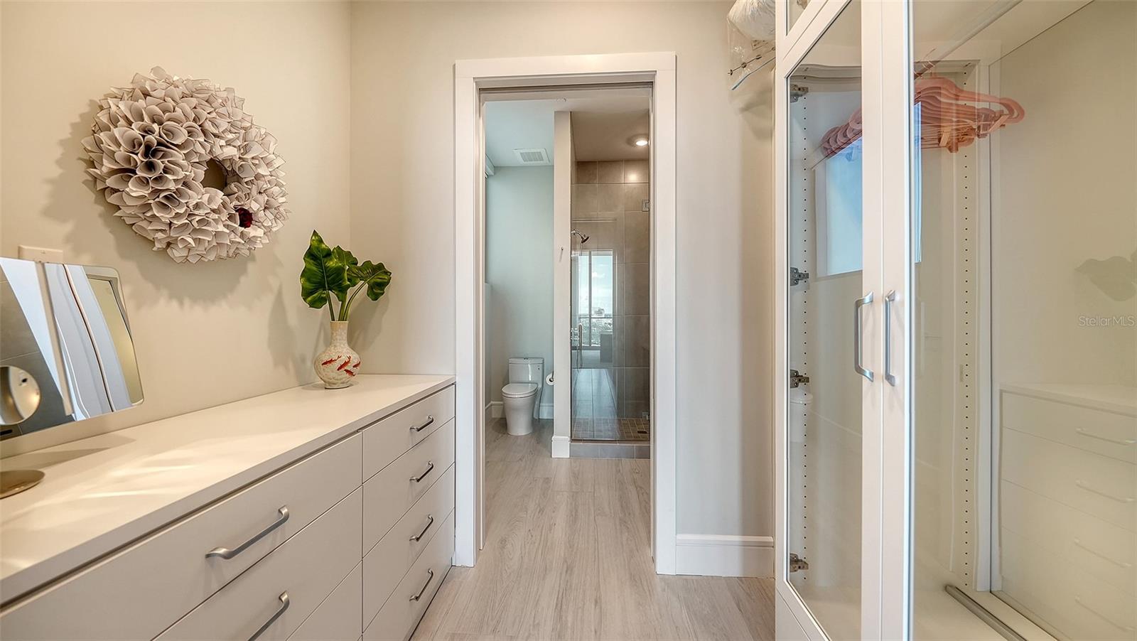 Dressing area with a long white vanity and glass-front closet beside an open bathroom doorway.
