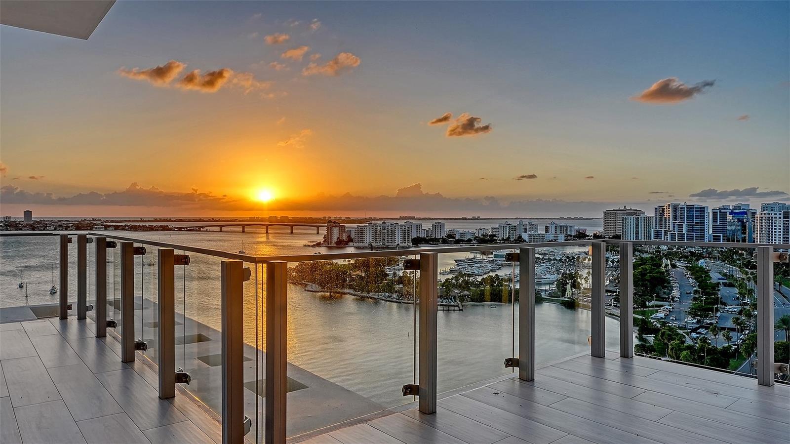 High-rise balcony with glass railings overlooking Sarasota Bay and the city at sunset.