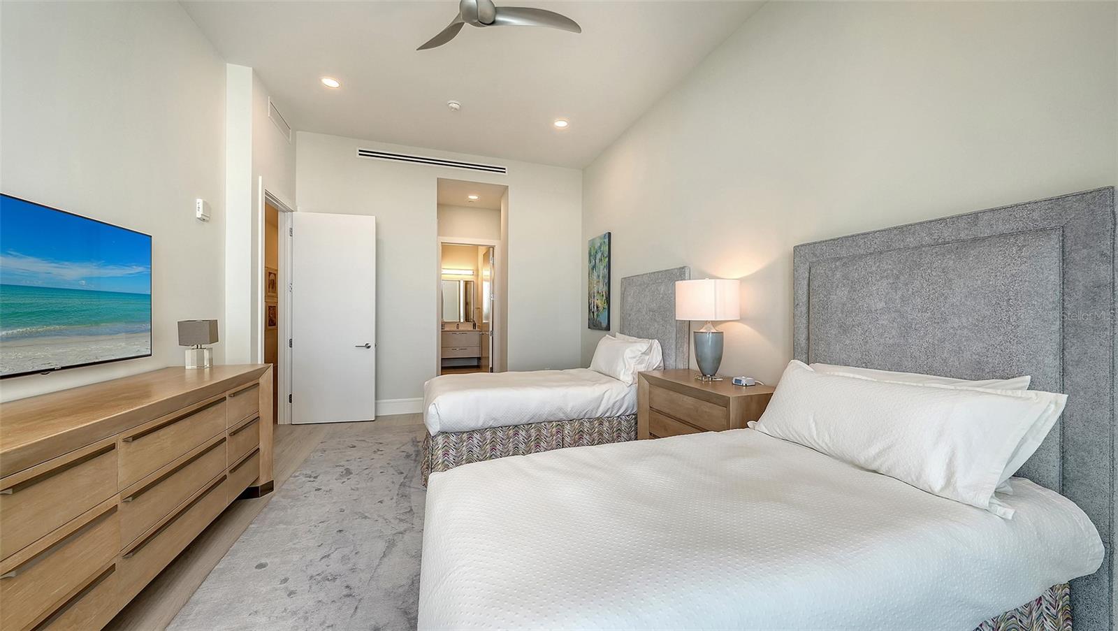 Bedroom with two white beds, gray upholstered headboards, and a television showing a beach.