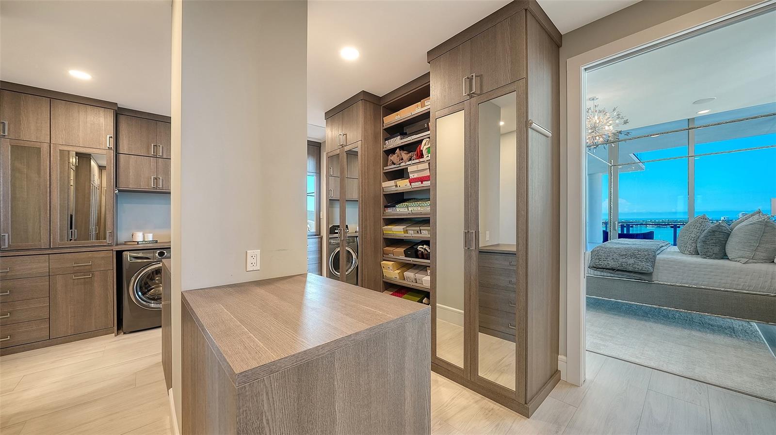 Walk-in closet with a central island, built-in laundry, and mirrored cabinetry beside a bedroom with a water view.