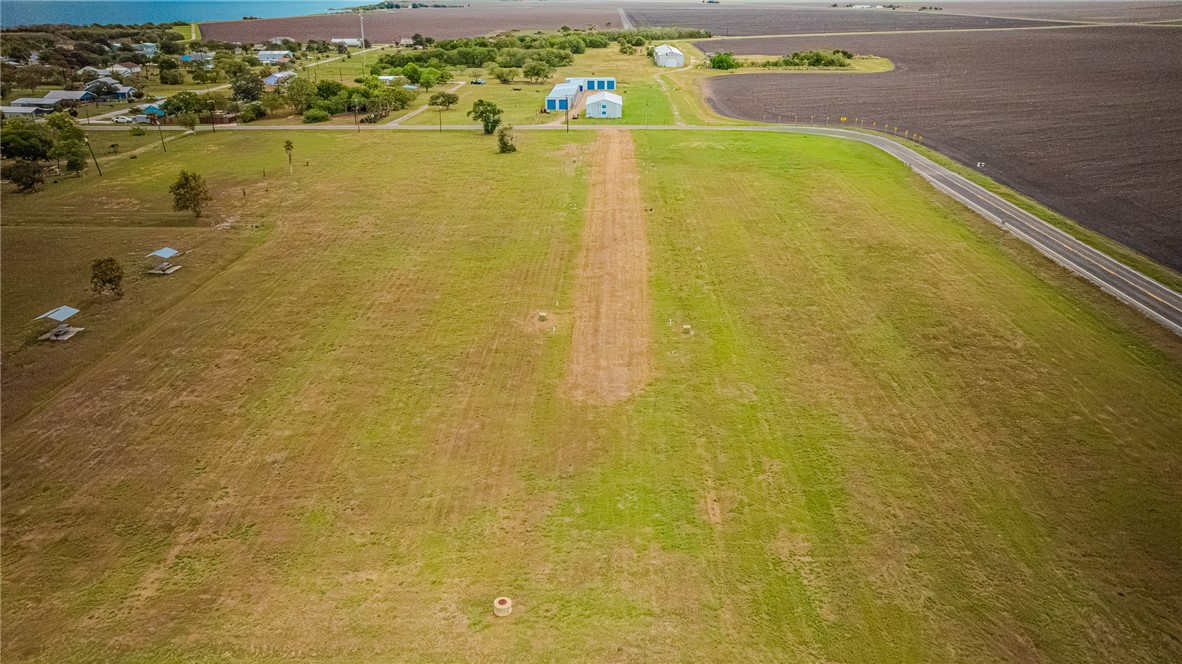 Aerial view of open grassy land with a dirt track, blue-roofed buildings, and a paved road.